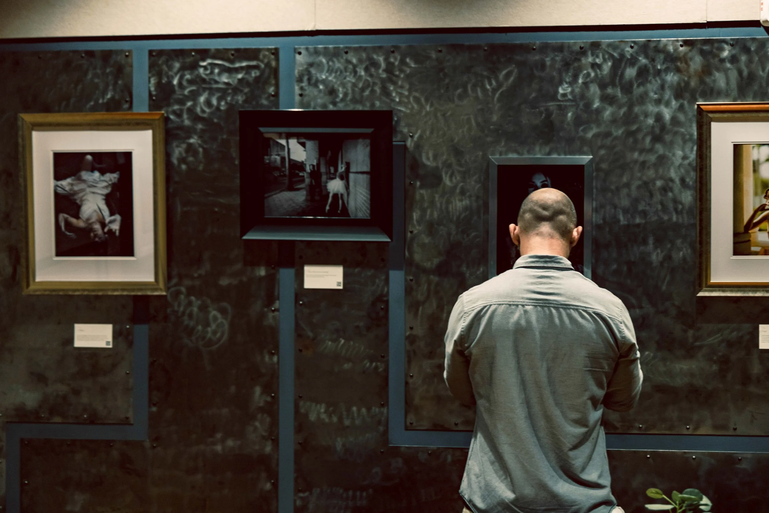 A person viewed from behind stands in an art gallery, looking at framed photographs on a dark textured wall.