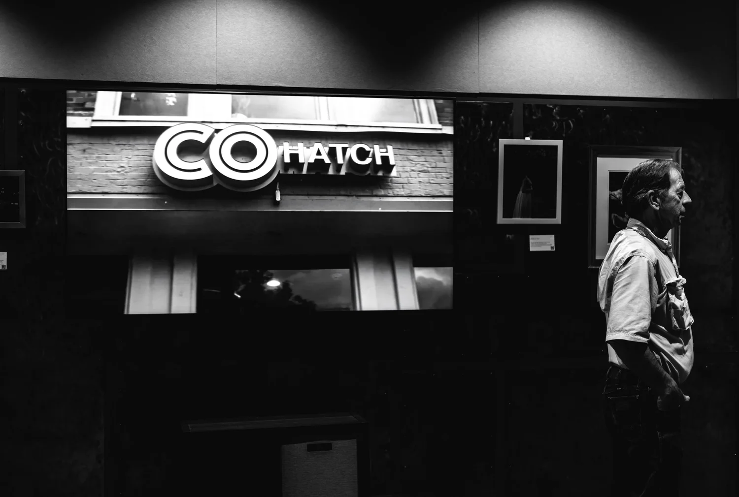 A man stands in profile in front of a dark wall with framed pictures, looking to the right. To the left, a screen displays an image of a sign that reads 'COHATCH' in front of a brick building.