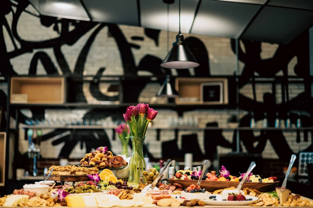 A table filled with various desserts, fruits, and snacks, with a tall glass vase of pink tulips in the center, set against a wall with abstract black and white graffiti art.