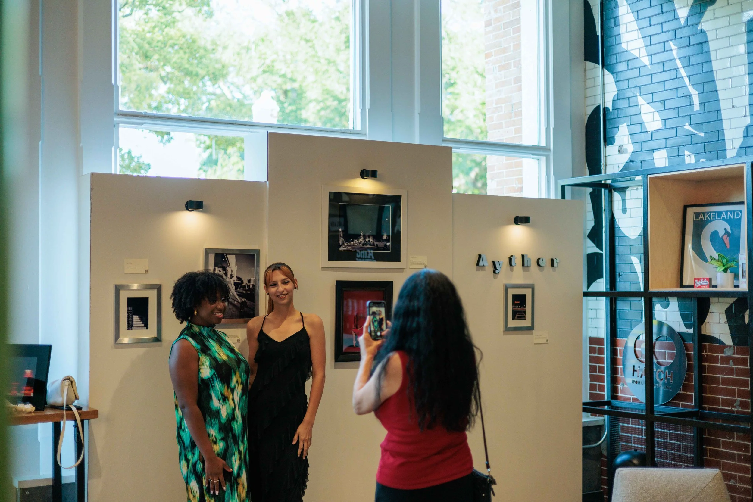 A woman in a red top taking a photo of two women posing in an art gallery with artwork and signs on the wall, and large windows behind them