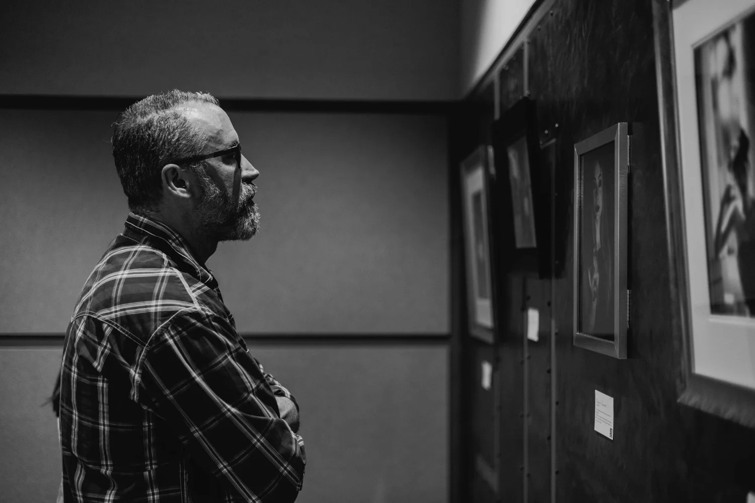 A man with glasses, a beard, and a plaid shirt, standing in profile in an art gallery, observing framed artwork on a wall.