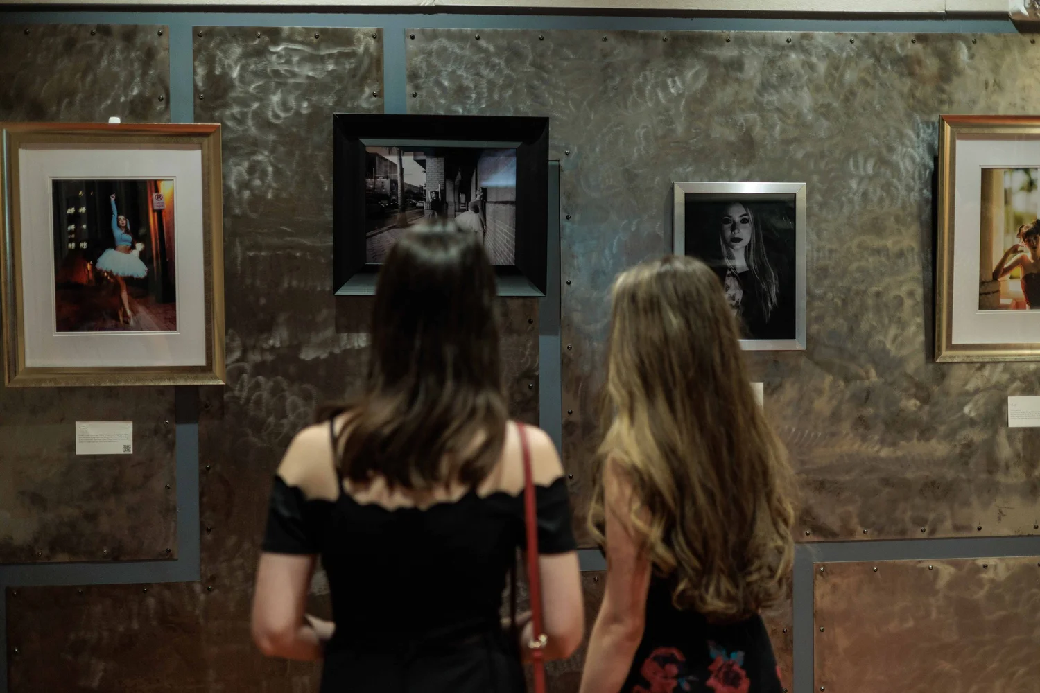 Two women observe photographs displayed on a textured metal wall in an art gallery.