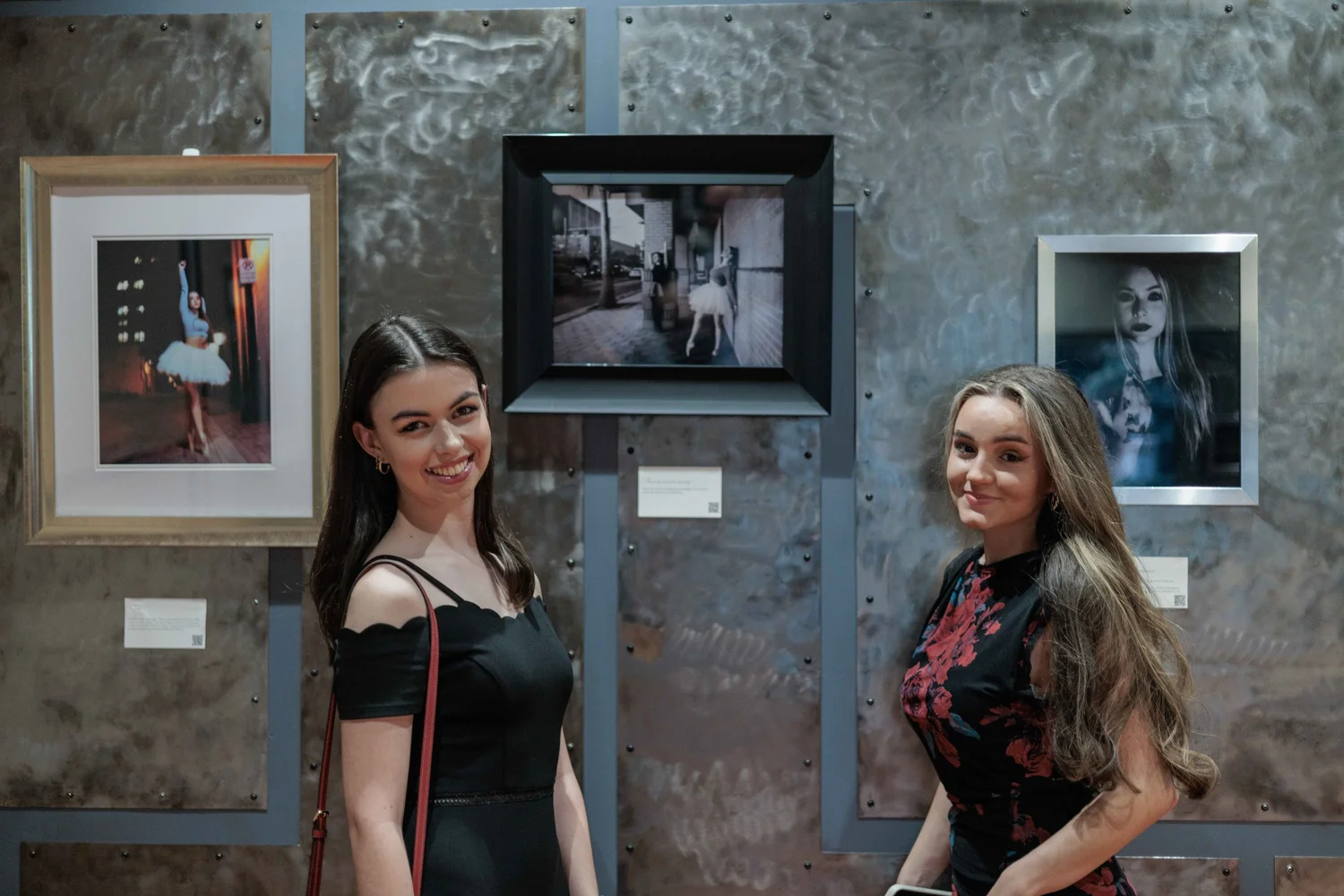 Two women posing at an art gallery with framed photographs on a textured wall behind them.