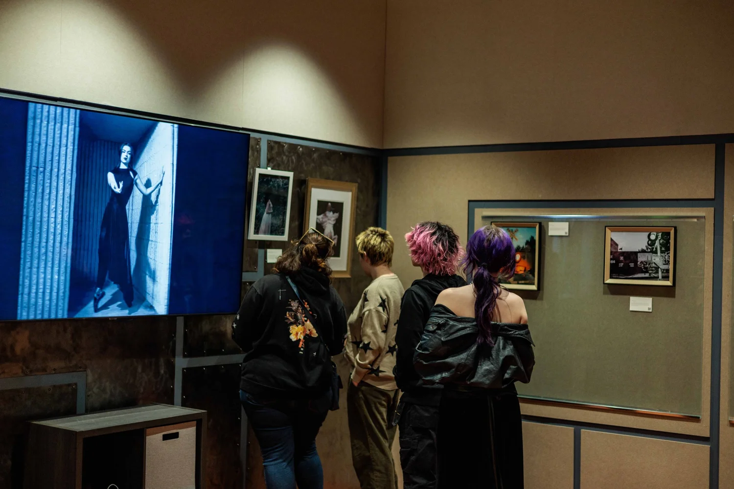 People viewing artwork in an art gallery, with framed pictures and a large blue-toned photo on a screen.