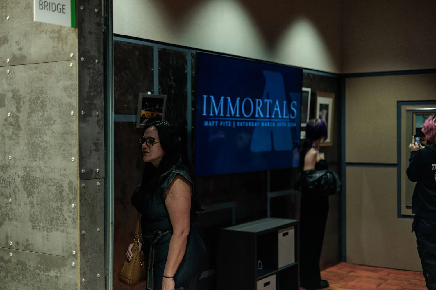 People at an event in a hallway with a large screen reading "IMMORTALS" and details about a performance by Matt Fitz, Saturday March 30th, 2024.