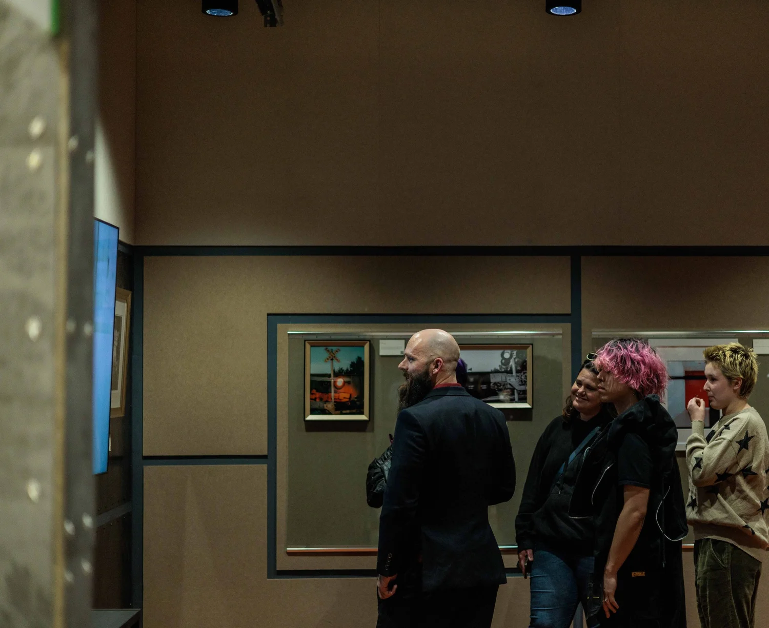Group of four people inside an art gallery, looking at paintings on the wall. A man with a bald head and beard is in profile, three women, one with pink hair, are smiling, and a young woman in a beige sweater stands on the right.