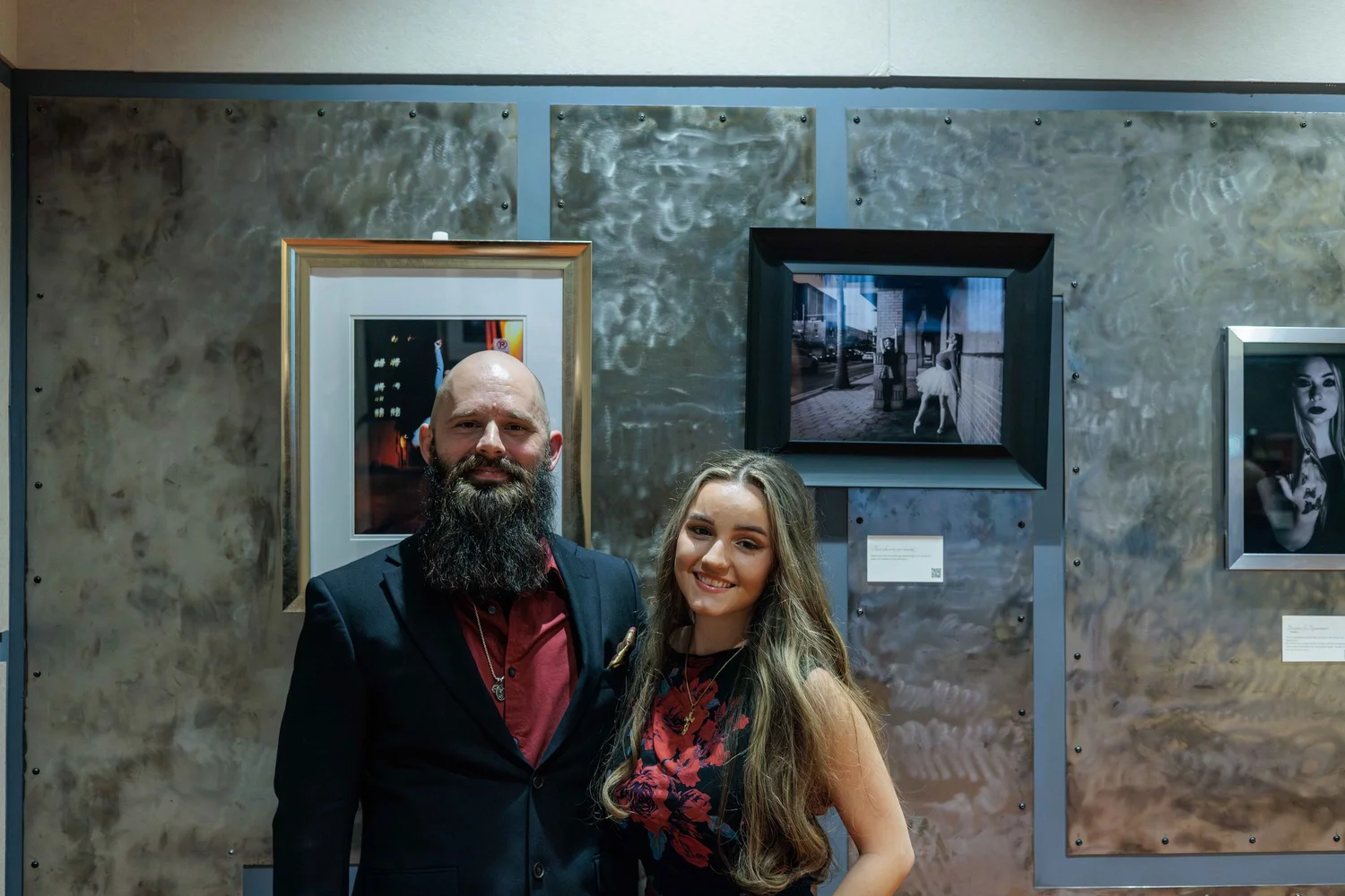 A man with a beard and a woman with long hair smiling, standing in front of a metallic wall with framed photographs at an art gallery.