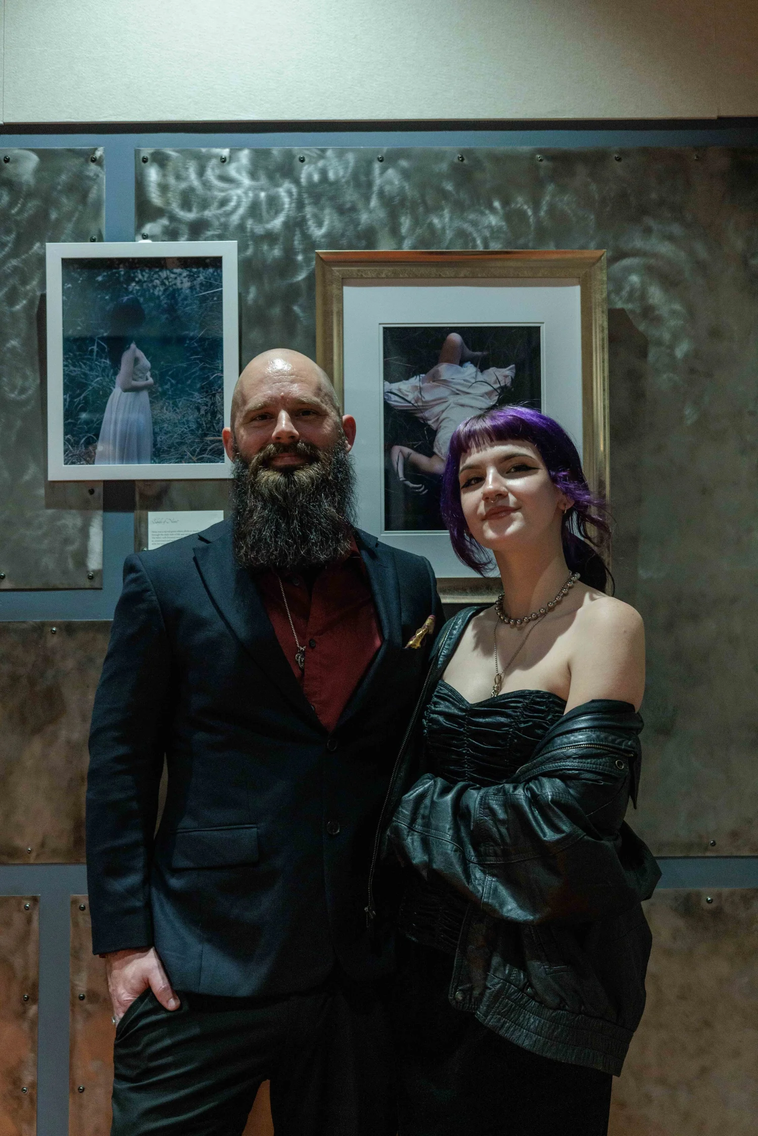 A man with a beard in a black suit and a woman with purple hair and black outfit posing together at an art gallery.