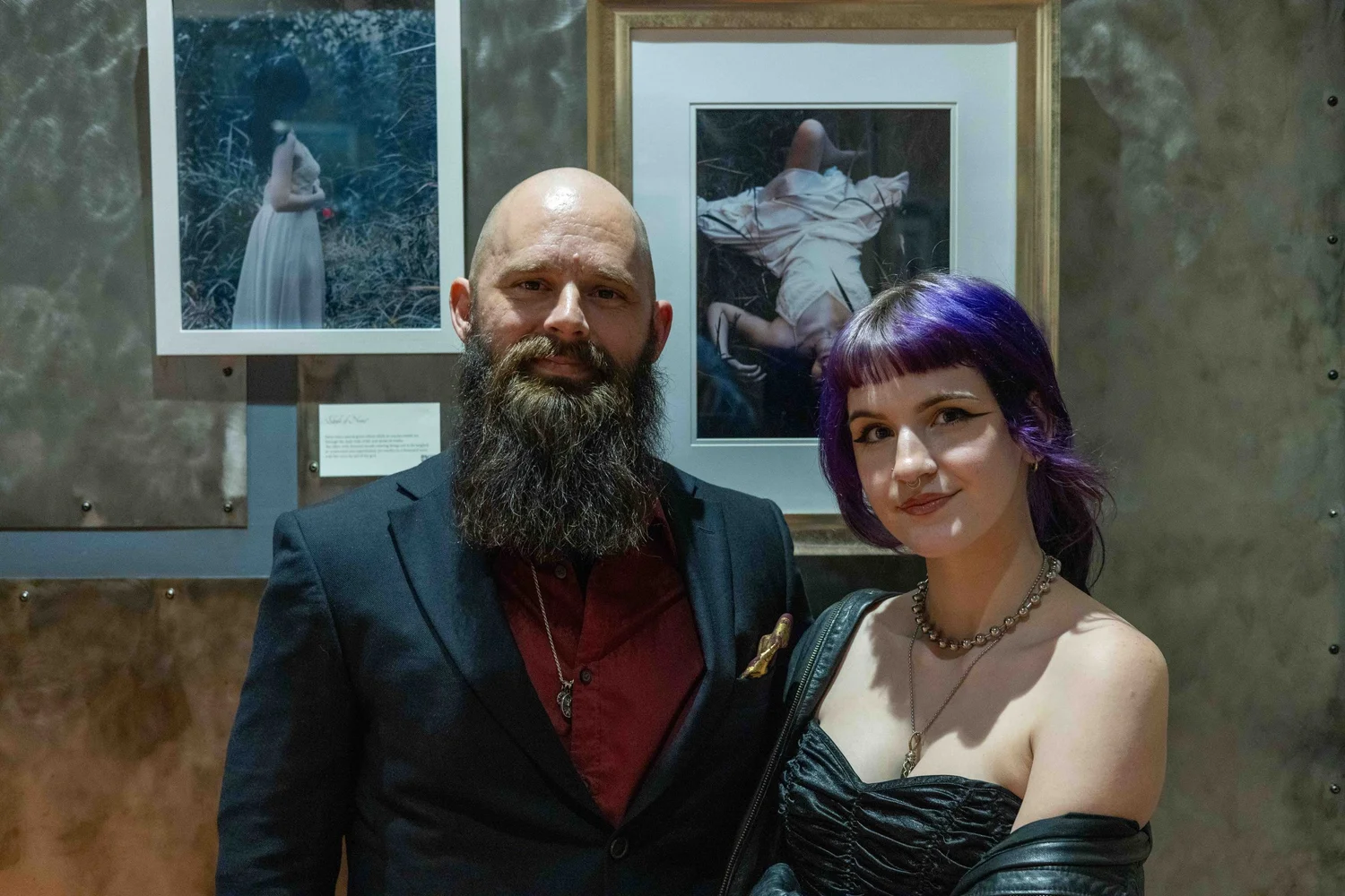 A man with a beard in a dark suit and a woman with purple hair in a strapless black dress standing close together in front of framed artwork at an art gallery.