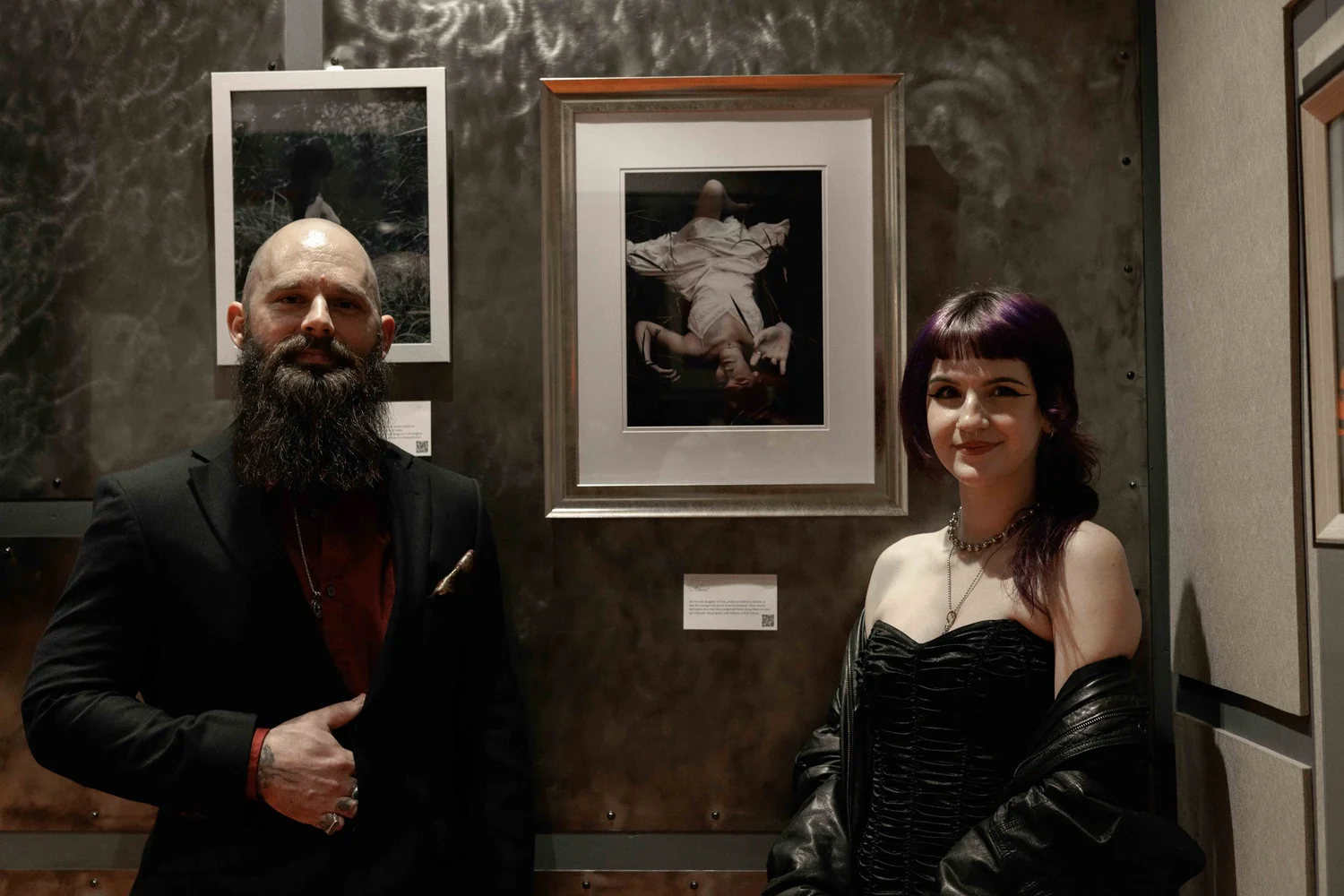 A man with a beard in a black suit and a woman with dark hair in a black dress posing in an art gallery in front of framed photographs featuring a woman lying down and a person sitting outdoors.
