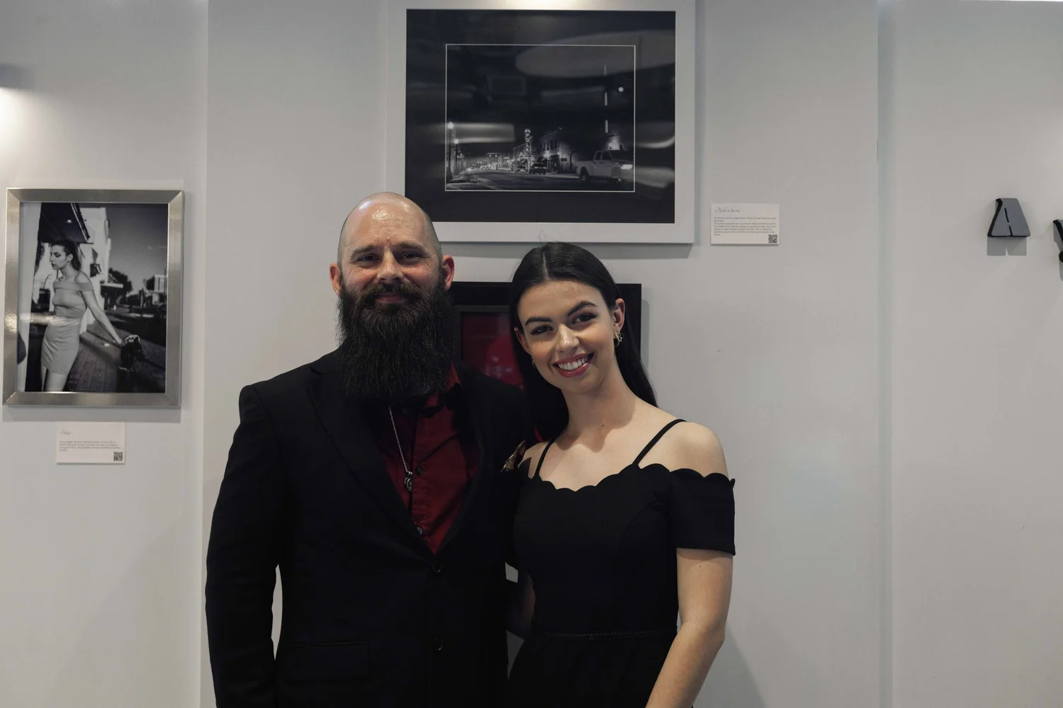 A man with a long beard and a woman with dark hair smiling, standing together at an art gallery. The man is dressed in a black suit with a red shirt, and the woman in a black off-shoulder dress. Behind them are framed black-and-white photographs on a