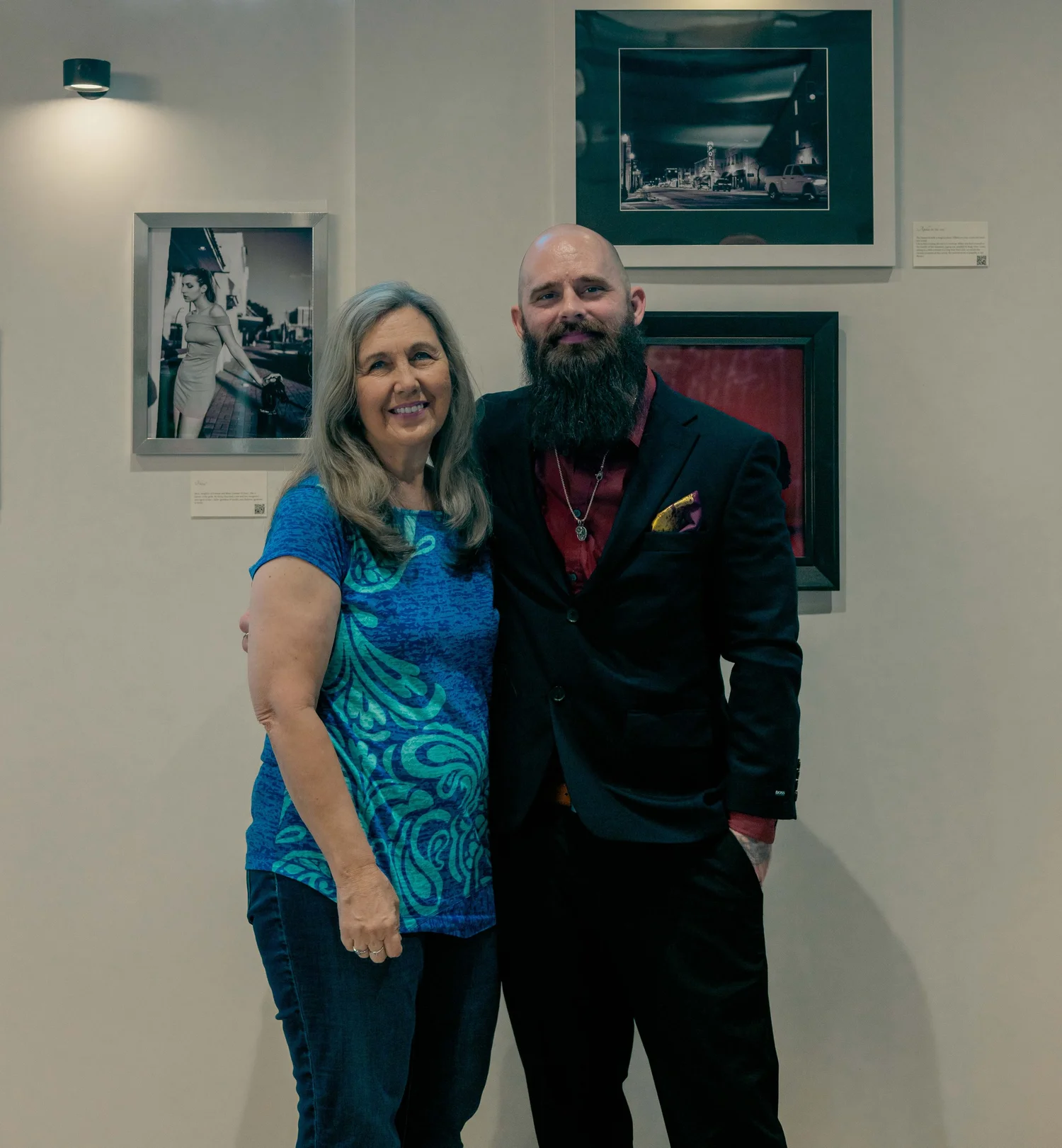 A woman and a man standing together, smiling for a photo in an art gallery. The woman has long gray hair and is wearing a blue patterned top. The man has a bald head, a long beard, and is dressed in a dark suit with a red shirt and a pocket square.