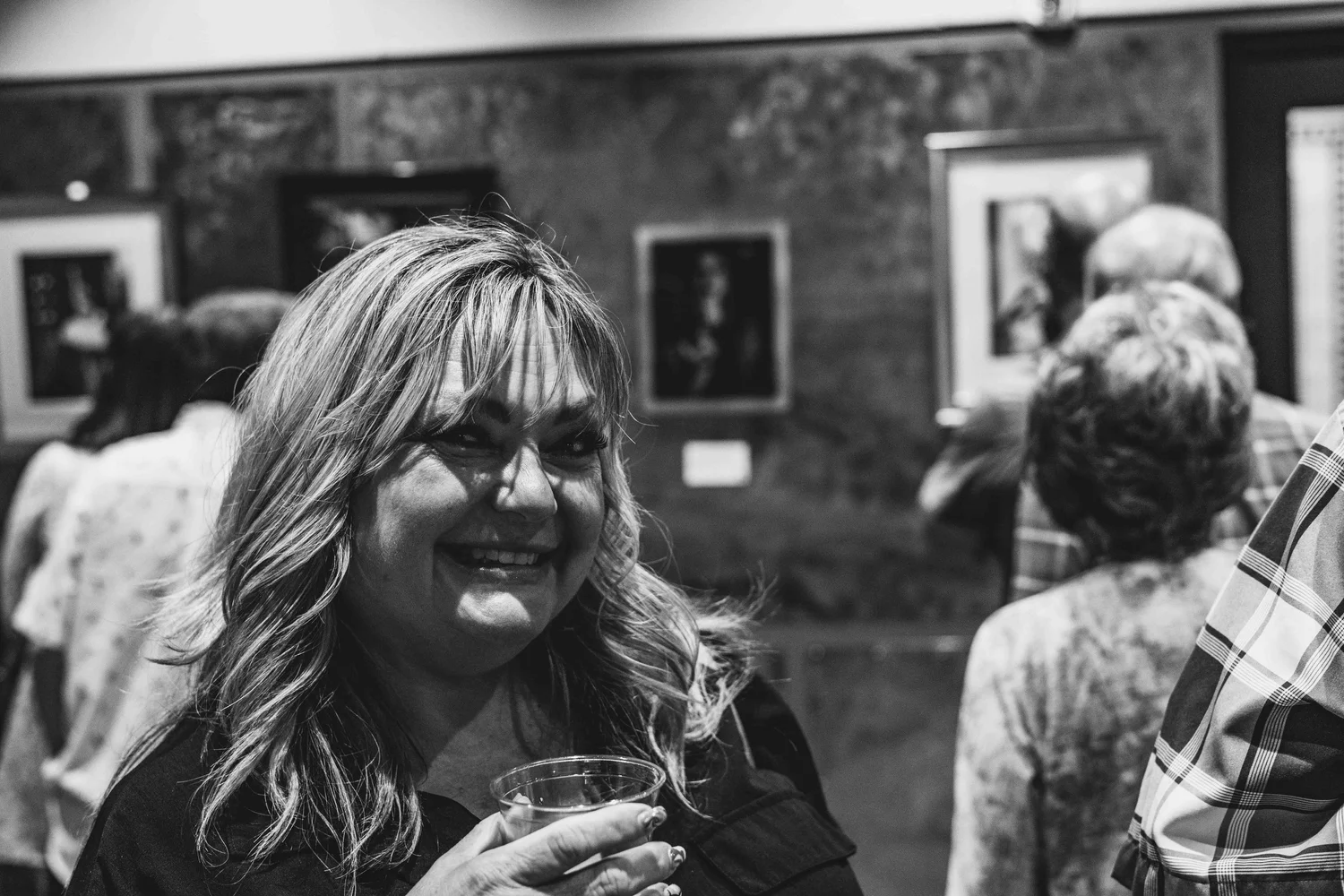 A woman with wavy hair smiling and holding a drink at an indoor event or social gathering, with other people and artwork on the wall in the background.