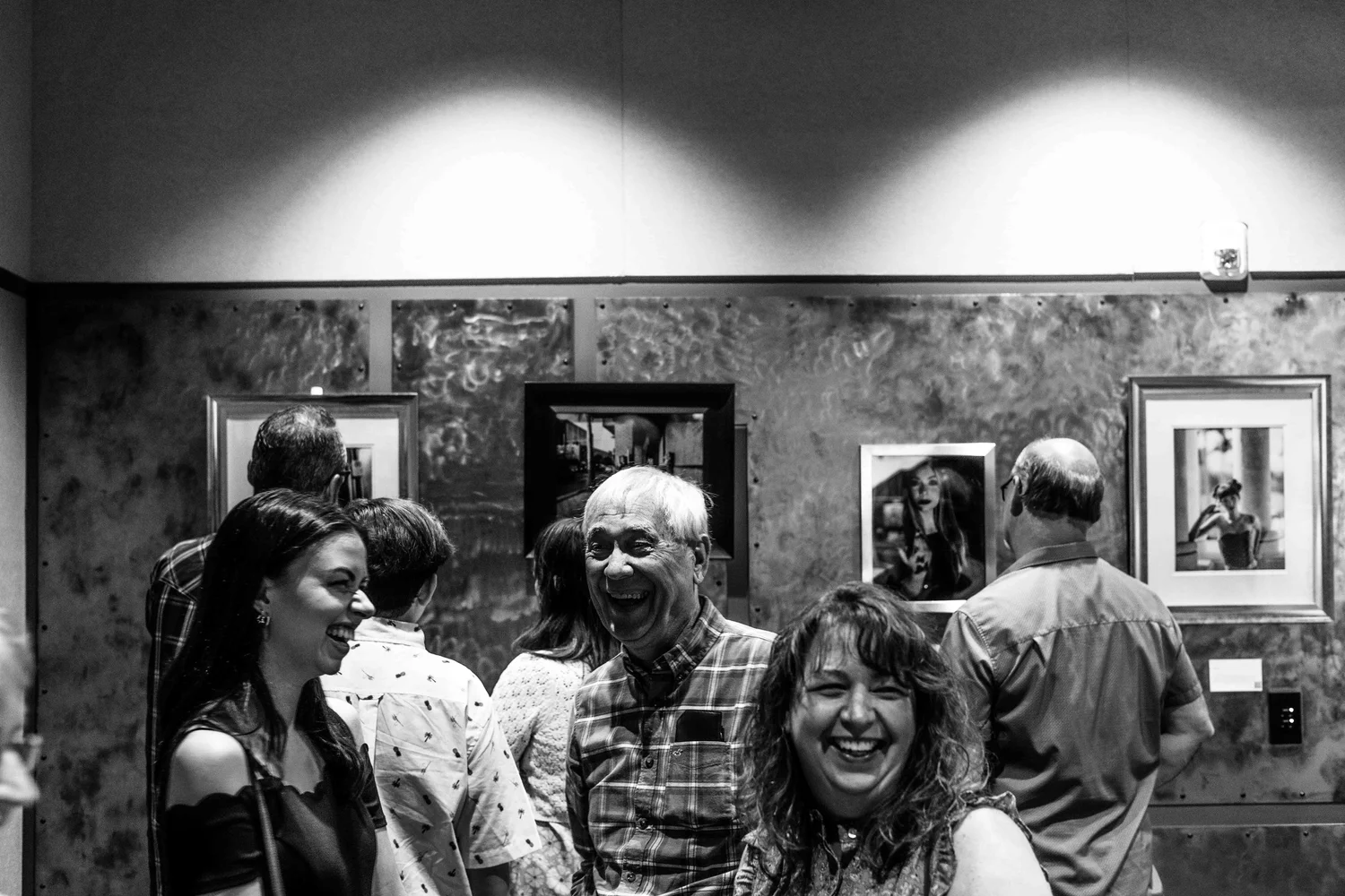 People smiling and laughing at an art gallery with framed photographs on the wall.