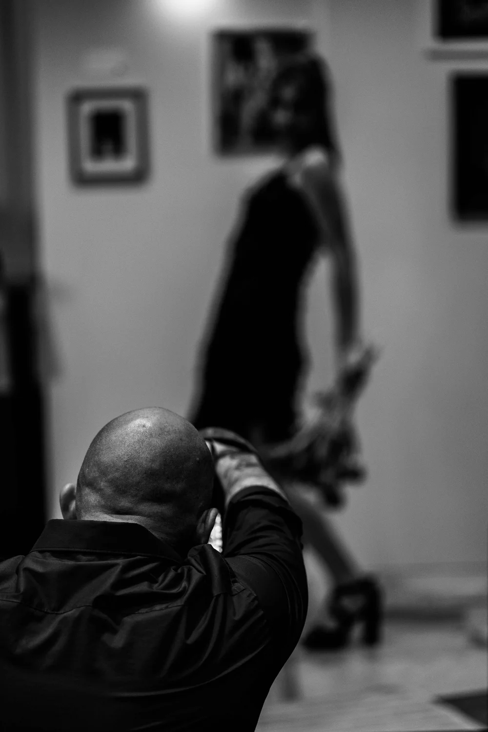 A man with a shaved head taking a photograph or video with a camera, with a blurred woman in the background in a room with framed pictures on the wall.